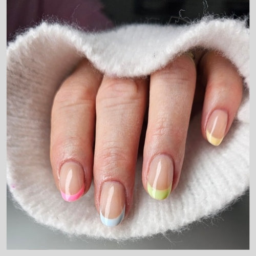 Een close-up van een hand met een stijlvolle French manicure waarbij elke nagel een andere kleur rand heeft. De gebruikte ShidoNails® kleuren zijn zachtroze (Bolero), lichtblauw (Gellak Mess), pastelgeel (Cocktail Pina Colada) en frisgroen (Nikki). De nagels hebben een natuurlijke glanzende basis en de hand rust in de mouw van een witte gebreide trui.